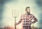 Young bearded farmer in red checkered shirt with old pitchfork sky nature backgrund, toned