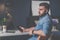 Young bearded freelancer man in denim shirt is sitting at home at table, holding mobile phone in his hand and thinking.