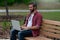 A young bearded hipster man button down red shirt sits on a park bench outside on a cloudy covered day near a walking path working