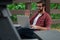 A young bearded hipster man with glasses sits against a pavilion pillar outside on a cloudy covered day near a walking path