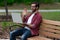 A young bearded hipster man with glasses sits on a park bench outside on a cloudy covered day near a walking path working remotely