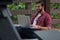 A young bearded hipster man sits against a pavilion pillar outside on a cloudy covered day near a walking path working remotely on