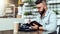 Young bearded hipster man sits in cafe at table, writes in notebook. On table closed laptop, cup of coffee, camera.