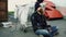 Young bearded homeless man sitting on a sidewalk near shopping cart ang garbage container during cold winter day