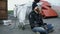 Young bearded homeless man sitting on a sidewalk near shopping cart ang garbage container during cold winter day