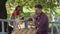 Young bearded lazy man drinking tea in the foreground while angry attractive girl in retro clothes planting flowers in