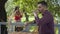 Young bearded man drinking tea in the foreground while attractive girl in retro clothes planting flowers in pots in the