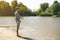 Young bearded man fishing standing on the sandy river bank with fish-rod during summer d