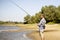 Young bearded man fishing standing on the sandy river bank with fish-rod during summer d