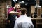Young bearded man getting haircut by hairdresser with electric razor at barber shop