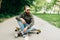 Young bearded man longboarder in casual clothes sitting on the longboard or skateboard outdoors, side view. Urban, subculture,