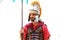 A young bearded man in a red uniform of a Roman legionary, in a metal helmet, tunic and with a spear stands in