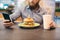 Young bearded man in shirt sitting in cafe with burger and paper cup holding smartphone online