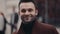 Young bearded man stands in the crowded street and smiles towards the camera. Stylish outfit, modern male fashion