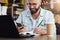 Young bearded man trendy glasses sits cafe in front of laptop computer, uses smartphone, takes notes in notebook