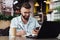 Young bearded man trendy glasses sits cafe in front of laptop computer, uses smartphone, takes notes in notebook