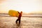 Young bearded surfer in wetsuit with yellow surfing longboard walk from water at sunset ocean. Water sport adventure camp and