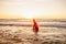 Young bearded surfer in wetsuit with yellow surfing longboard in a water at sunset ocean. Water sport adventure camp and extreme