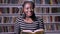 Young beautiful african american woman reading book in library, smiling, happy, bookshelves background