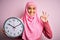 Young beautiful arab woman wearing pink muslim hijab doing countdown holding big clock doing ok sign with fingers, excellent