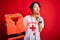 Young beautiful asian lifeguard girl using whistle holding orange life jacket over red background serious face thinking about