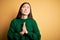Young beautiful asian woman wearing green winter sweater over yellow  background begging and praying with hands together