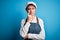 Young beautiful baker woman with blue eyes wearing apron and cap over blue background with hand on chin thinking about question,