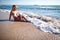 Young beautiful blond woman in white bikini sitting on sea water edge and enjoying sunshine on clear summer day