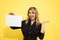 Young beautiful blonde woman in black dress holds empty sheet for inscription on yellow background in hands pointing with hand,