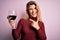 Young beautiful blonde woman drinking glasse of red wine over isolated pink background very happy pointing with hand and finger