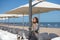 Young beautiful blonde woman leaning on a lamppost on the promenade next to the white umbrellas and chairs on a terrace. On the