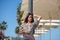 Young beautiful blonde woman leaning on a lamppost on the promenade next to the white umbrellas and chairs on a terrace. In the