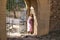 Young, beautiful, blonde woman in a pink dress, leaning against the wall of the entrance of a castle receiving the sun\'s rays