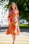 Young beautiful blonde woman in a red flower dress crosses the road at a crosswalk