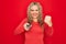 Young beautiful blonde woman using television remote control over isolated red background annoyed and frustrated shouting with