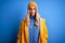 Young beautiful blonde woman wearing raincoat for rainy weather over blue background with serious expression on face