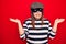 Young beautiful brunette burglar woman wearing cap and mask over isolated red background clueless and confused with open arms, no
