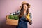 Young beautiful brunette farmer woman wearing apron and hat holding box with plants with surprise face pointing finger to himself