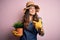 Young beautiful brunette farmer woman wearing apron and hat holding pot with plants with surprise face pointing finger to himself