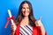 Young beautiful brunette smart woman holding graduated degree diploma over blue background smiling happy and positive, thumb up