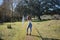 Young, beautiful, brunette woman with curly hair, white top, jeans and heels, standing looking at camera in the middle of a park