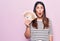 Young beautiful brunette woman holding euros banknotes over isolated pink background scared and amazed with open mouth for