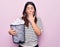 Young beautiful brunette woman holding full paper bin of crumpled papers over pink background covering mouth with hand, shocked