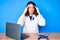 Young beautiful brunette woman wearing doctor uniform with blue ribbon with hand on head, headache because stress