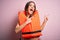 Young beautiful brunette woman wearing orange lifejacket over isolated pink background smiling and looking at the camera pointing