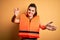 Young beautiful brunette woman wearing orange safe lifejacket over yellow background looking at the camera smiling with open arms