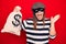 Young beautiful burglar woman wearing cap and mask holding money bag with dollar symbol celebrating achievement with happy smile