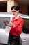 Young beautiful business woman with red lipstick in glasses and red polka dots blouse standing by her luxurious car