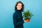 Young and beautiful caucasian girl with curly hair keeps green flower in a pot and smiles, portrait isolated on blue