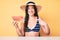 Young beautiful caucasian woman wearing bikini and hat holding slice of watermelon pointing finger to one self smiling happy and